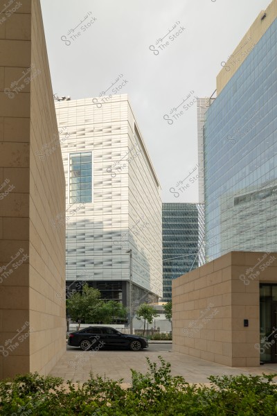 An urban scene featuring several modern buildings with glass and concrete facades. A black car is parked on the street between the buildings. The sky is overcast, and some green trees add a touch of nature to the urban area.