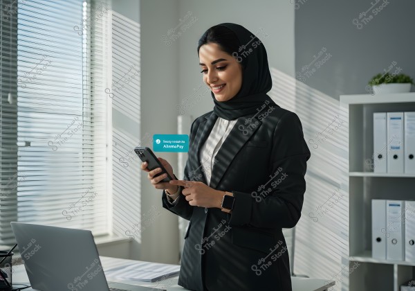 An image of a woman wearing a hijab and a black formal suit standing in a well-lit office near a window with venetian blinds. She is holding a smartphone with a smile, looking at it, and wearing a smartwatch. In the background, there are shelves with organized files and a decorative plant. The \"AlinmaPay\" logo is visible in the image.