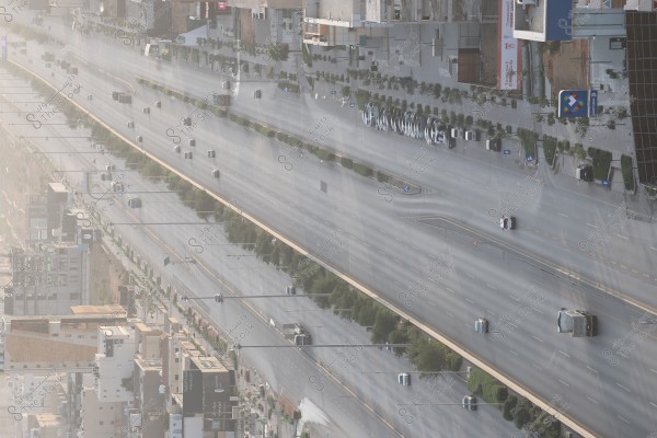 صورة جوية لشارع رئيسي مزدحم في مدينة, تظهر المركبات المتحركة في كلا الاتجاهين مع وجود أشجار على جانبي الطريق. المباني واللافتات التجارية تظهر على جانبي الشارع, مشيرة إلى منطقة حضرية حديثة واسعة. الإضاءة تشير إلى أن الصورة قد تكون ملتقطة في وقت مبكر من النهار أو عند الغروب.\r\n\r\n####