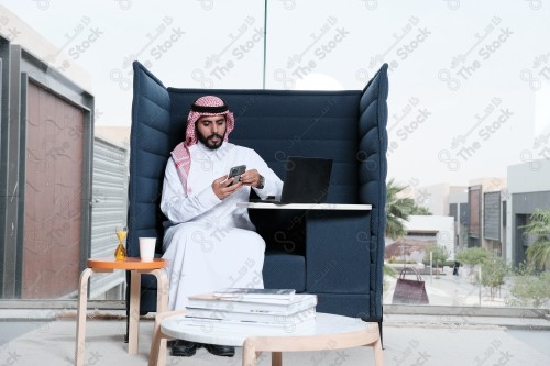 رجل سعودي يرتدي الزي السعودي التقليدي يقوم بإجراء مكالمة هاتفية بينما يدون ملاحظاته على إحدى صفحات المفكرة ويظهر الجهاز المحمول على سطح الطاولة أثناء النهار