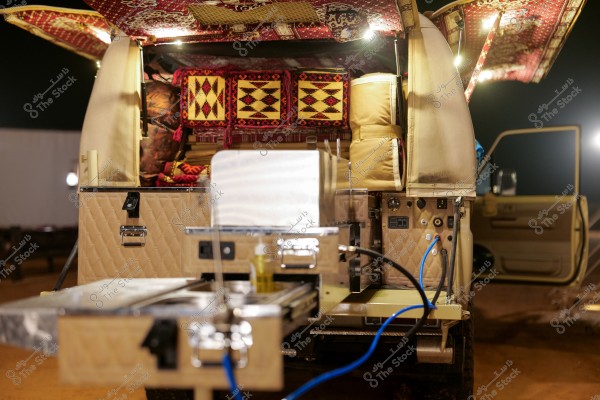 The image shows the back of a vehicle equipped for camping or desert trips. The vehicle has cushioned, open cabinets with kitchen equipment such as a stove and drawers, connected with electrical wires. The top of the vehicle is lined with traditional rugs, and above the cabinets are cushions decorated with traditional Arabic patterns. The vehicle is parked in a desert-like environment at night.