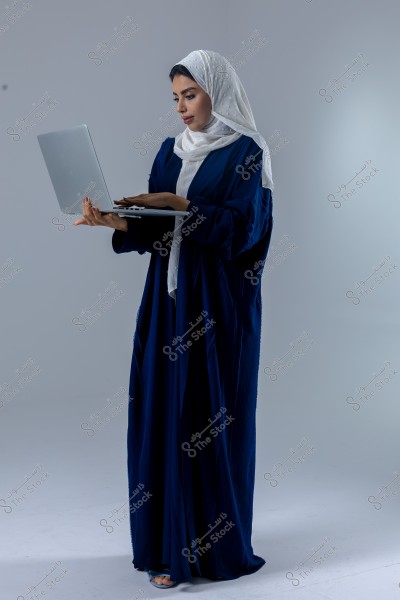 A woman wearing a dark blue abaya and a white hijab is holding a laptop. She stands in a side pose, looking at the laptop screen. The background is light gray.