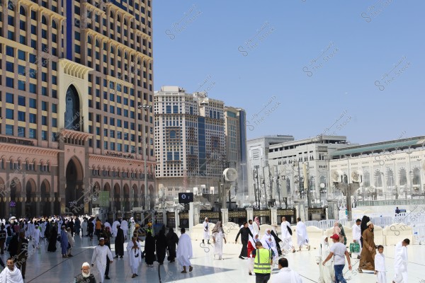 The image shows a large open courtyard in Saudi Arabia crowded with people wearing white Ihram garments, possibly in Mecca near the Grand Mosque. In the background, there are tall buildings with modern architectural facades and Islamic motifs.