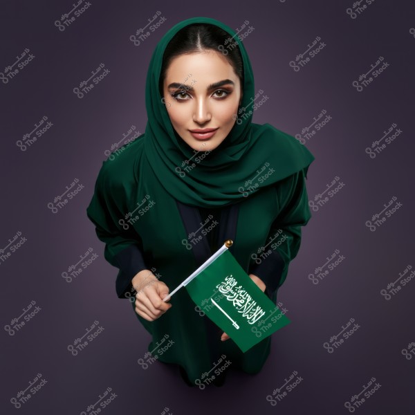 A portrait of a woman wearing a green abaya and a dark green hijab, holding a Saudi Arabian flag. The photo is taken from an overhead angle, and the woman is gently smiling at the camera. The dark background highlights the details of the clothing and flag.