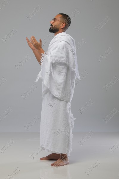 The image shows a man wearing white Ihram clothing, standing in a prayer position with his hands raised. He is wearing glasses and has a short beard. The setting appears to be indoors with a simple gray background.