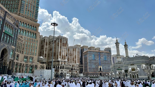 مشهد للحرم المكي الشريف في مكة المكرمة, يظهر فيه الحجاج والمعتمرين بملابس الإحرام, مع خلفية من الأبراج العالية والسماء الزرقاء المزينة بالغيوم.