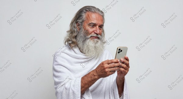 A portrait of an elderly man with a long white beard and hair holding a smartphone. He is wearing a white garment resembling an Ihram. The background is white.