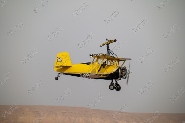 A vintage yellow biplane flying in the air, with the number \"2\" visible on the tail. A person wearing a flight suit is standing on a structure above the plane. The background shows a clear sky and some desert terrain.