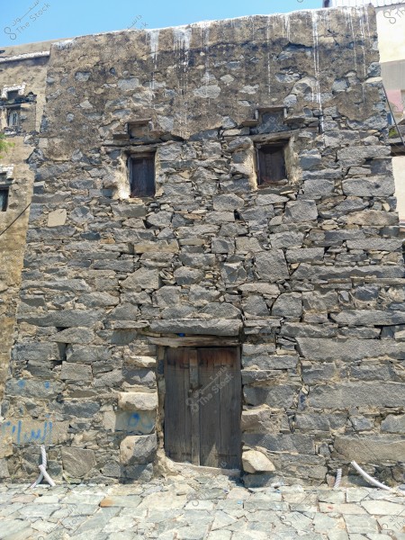 Image of an old building made of dark gray stone with brown hues. The facade is rectangular with two small wooden windows at the top and a wooden door at the bottom. The roof appears slightly weathered, and there are some blue writings on the wall. The ground is covered with large similar stones.