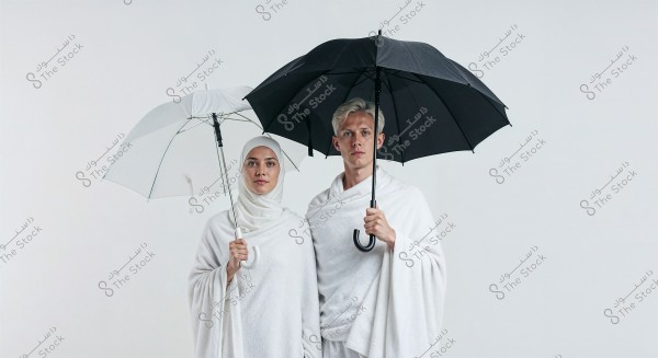 A photo of a woman and a man standing together, both wearing white Ihram clothing, which is traditionally worn by Muslims during Hajj and Umrah. The woman is wearing a hijab and holding a white umbrella, while the man holds a black umbrella. The background is a simple white.