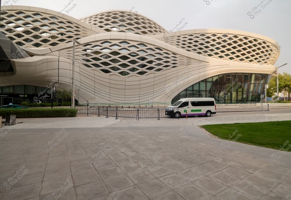 A modern architectural building set in an urban complex, showcasing an advanced design with undulating beige latticework and glass that reflects sunlight. A small bus in white and green colors is parked beside the building, with an open paved space and green grass in the foreground.