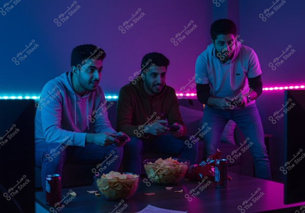The image shows three young men sitting in a dimly lit room glowing with blue and purple neon lights. They are focused on a screen in front of them, holding video game controllers. The young men are dressed casually, mostly in hoodies and jeans. On the table in front of them, there are bowls filled with potato chips and soda cans. The atmosphere appears lively and exciting.