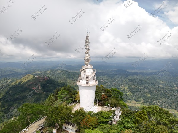 An image of a tall white tower situated on a plateau covered with dense green trees, with scenic mountains and thick clouds forming the sky in the background. The tower features a unique spiral design at the top.