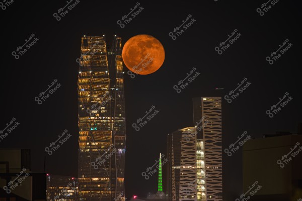 A nighttime scene of illuminated skyscrapers in a city, with a large orange moon in the dark sky behind them. The buildings feature modern and intricate architectural designs, with a green light visible below the moon.
