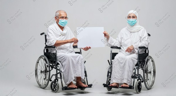  The image shows two elderly people sitting in wheelchairs. Both are dressed in white ihram clothing and wearing medical face masks. They are holding a blank white sign. The attire suggests they may be pilgrims on a religious journey such as Hajj or Umrah.