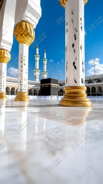 Image of the Kaaba in Mecca, Saudi Arabia. The foreground features elaborately decorated pillars with gold and white designs, while two minarets are visible in the background under a clear blue sky. The marble floor reflects the surrounding light of the Kaaba.