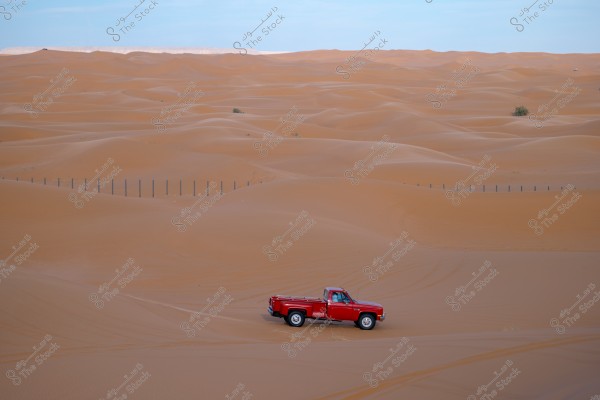 An image of a striking red truck situated in the midst of a desert with smooth, undulating sand dunes. The sky is clear in the background, while a row of posts stretches across the horizon, adding an element of structure to the expansive landscape of rolling sands.