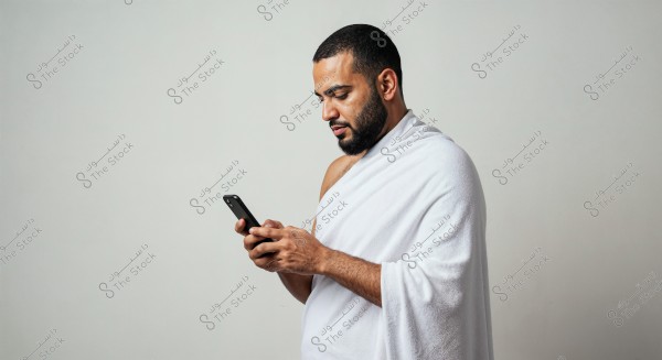 A portrait of a man wearing a white Ihram, focused on his mobile phone. The background is neutral and the atmosphere is calm, highlighting his facial features and concentration on the phone.