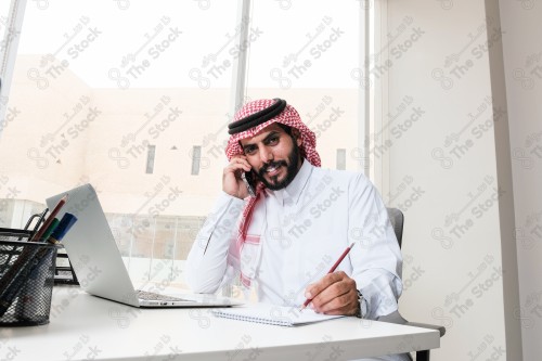 رجل سعودي يرتدي الزي السعودي التقليدي يعمل في مكتب بواجهة زجاجية ويستخدم قلم رصاص لتدوين الملاحظات