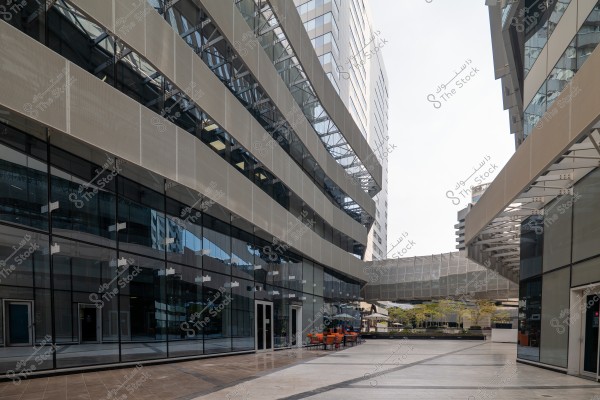 An image of a modern building facade featuring a glass and metal design with curved, multi-story architectural lines. The expansive pavement and walkways are visible in front of the building, with orange chairs and tables arranged alongside the glass walls on either side of the image. A metallic overhead bridge connects two parts of the building, and several small trees are visible in the background.