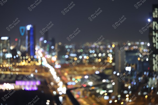 A nighttime cityscape with illuminated buildings and streets, appearing blurred. Tall buildings with lights are visible in the background, creating a bokeh effect that reflects the city lights.
