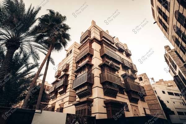 تُظهر الصورة مبنى تقليديًا بهندسة معمارية مميزة مع نوافذ مزخرفة من الخشب البني الداكن. المبنى محاط بأشجار نخيل شامخة تتمايل بلطف. السماء خلفية صافية مما يعطي للمشهد شعورًا بالهدوء والتراث. يبدو أن البناء يحتوي على عدة طوابق يُغطيها نمط معماري عربي قديم, وقد يكون من المباني التراثية في جدة, المملكة العربية السعودية.