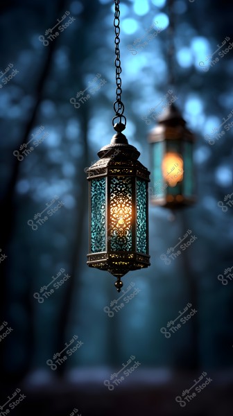 Two metallic lanterns with bright lights hang in the foreground of the image, featuring intricate designs that reflect Islamic motifs. The background is blurred, showing mysterious trees in a nighttime setting with a dim blue light.