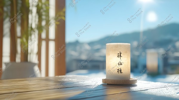 Image of a lit candle placed on a wooden surface, surrounded by a blurred outdoor landscape with mountains visible in the background under a blue sky. The candle is adorned with Japanese characters, and warm natural light is streaming in through the window.