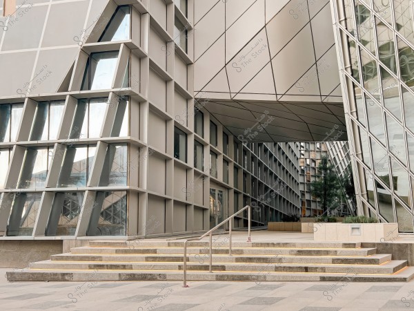 The image shows a modern architectural design featuring an angular structure with glass facades and metal frames, forming part of a functional and contemporary building. In the foreground, there are exterior stairs leading up to an entrance, and to the right, plants appear as part of the landscaping.