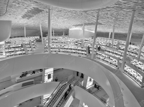 Image of a modern library with a unique architectural design, featuring multiple shelves filled with books and softly lit. The library includes circular escalators and a seating area. The ceiling has a distinctive geometric pattern. The image is in black and white and shows wide open spaces.