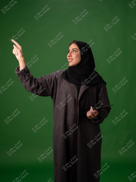 A portrait of a woman wearing a black abaya and hijab, standing and pointing upwards with her finger, while holding a tablet in her other hand. The background is green.
