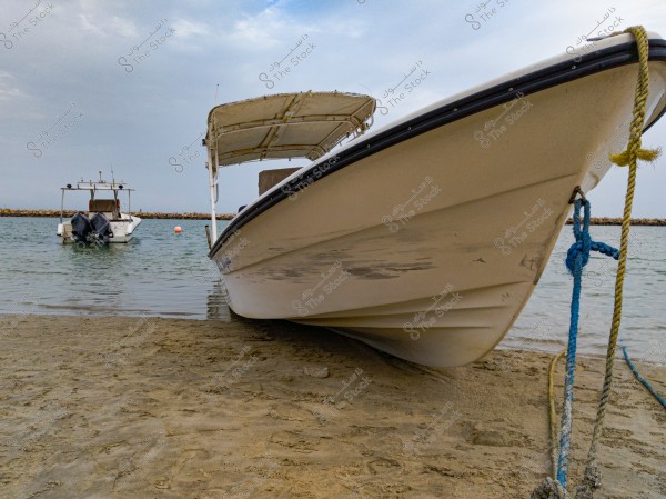 A large white boat is tied with a blue rope on the sandy shore, with calm water extending in the background. Another boat with two engines is seen in the water behind, and a cloudy sky is visible above.