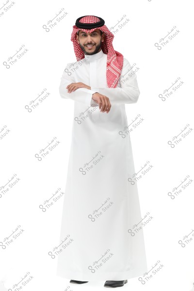 A portrait of a man wearing a traditional white thobe with a red checkered shemagh and a black agal, standing and smiling against a white background. The man has a confident appearance and seems to be from the Arabian Gulf region.