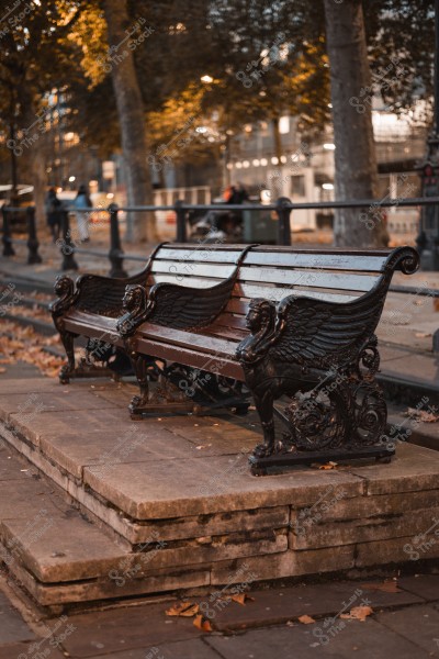 مقعد حديقة مزين بنقوش مزخرفة على جوانبه, محاط بأوراق الشجر المتساقطة وأشجار في الخلفية عند الغسق.