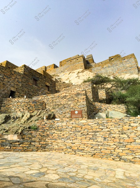 The image depicts a view of traditional architecture in an ancient village built from stones. The multi-level structures are tightly packed on a mountain slope under a blue sky. The stones vary in color between gray and golden brown, with a few green trees next to the buildings.