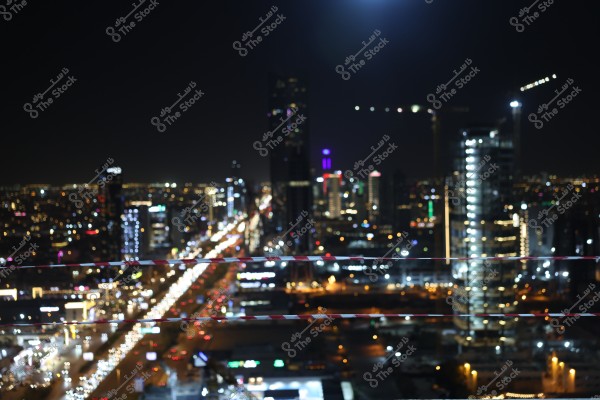 A blurry night view of a modern city adorned with lights and high-rise buildings. The lights are glowing on the streets and buildings in the background, with a dark sky above. Light trails of traffic can be seen on the roads.