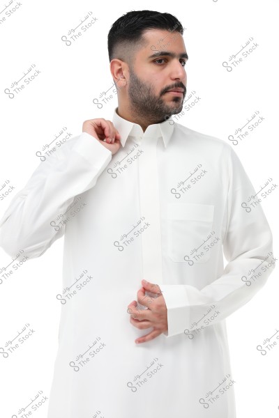 A man wearing a traditional white thawb, possibly of Gulf style. The man is positioned sideways, partially showing his facial features while adjusting the top part of his thawb with his hand. He has a neat beard and short, well-groomed hair. The background is white, emphasizing the details of the traditional attire.