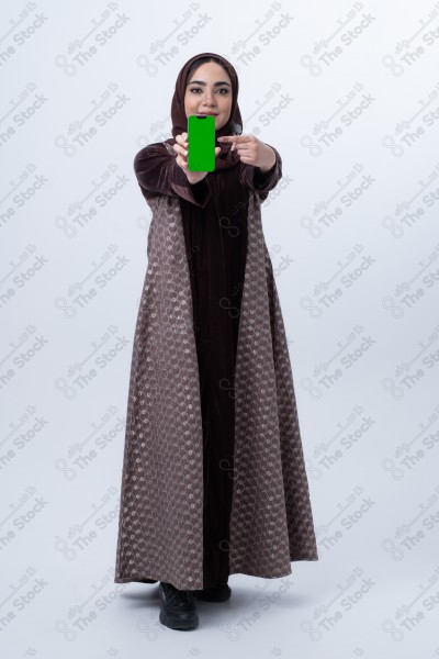 A young Saudi woman wearing an abaya and hijab, holding a green-screen phone