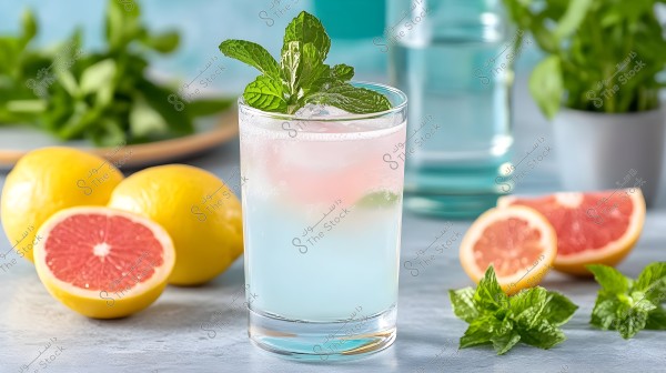 A glass cup filled with a bright blue beverage, topped with ice and fresh mint leaves. In the background, there are whole yellow lemons and a half grapefruit. In the distant background, a potted plant and some blurred green leaves are visible.