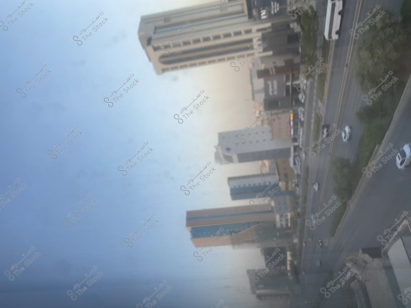 An image of a large city displaying several skyscrapers of various shapes and sizes, stretching along a wide street. Cars are moving on the street, with scattered trees on the side. The sky is clear and blue, indicating a sunny day.