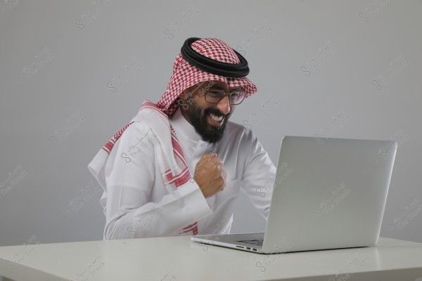 A man wearing traditional Saudi attire, including a thobe, ghutra, and agal, sits in front of a laptop on a desk. He appears to be smiling and excited, with his hand raised in a gesture of success or triumph.