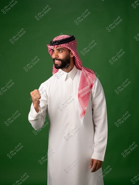 A portrait of a man wearing a white thobe and a red ghutra against a green background. The man is smiling in a half-body shot and raising his fist, as if expressing enthusiasm or pride. The red checked keffiyeh is neatly secured on the head with a black agal.