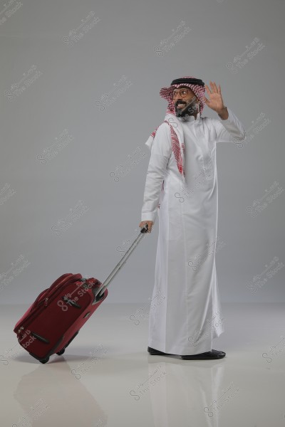 A man wearing a white thobe and a red checkered ghutra is pulling a red wheeled suitcase. He appears to be waving and has a smile on his face. The attire is traditional, reflecting Gulf culture, possibly from Saudi Arabia. The background is plain white.