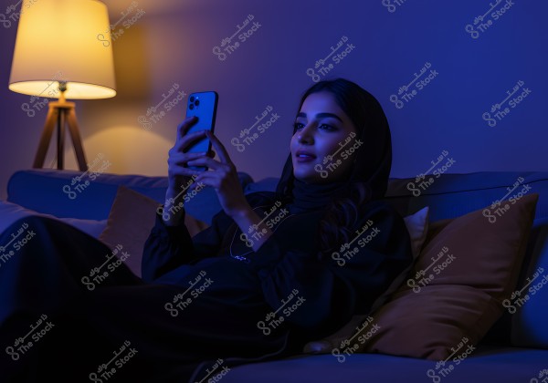 The image shows a woman sitting on a couch in a dimly lit room. She is wearing a black abaya and hijab, looking at a mobile phone in her hands with a calm smile. The room is lit by a floor lamp emitting warm light beside her, and cushions are placed around her.