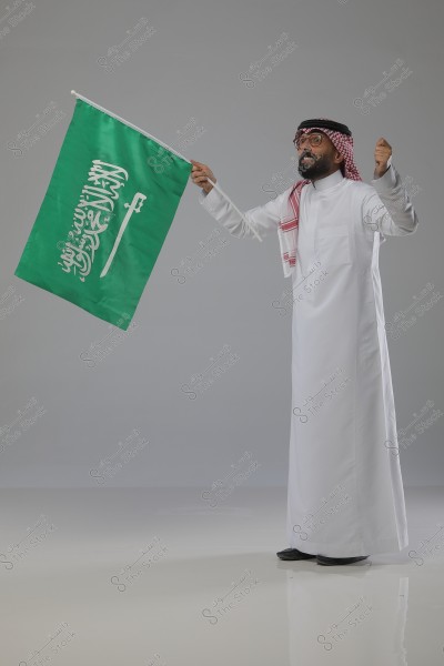An image of a man standing and holding the flag of Saudi Arabia. He is wearing a white thobe, a white and red ghutra with a black agal. He appears happy and is smiling against a gray background.
