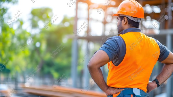 An image of a man at a construction site, wearing an orange hard hat and an orange safety vest. He is standing with his back to the camera, hands on hips, with blurred unfinished building structures and trees in the background.