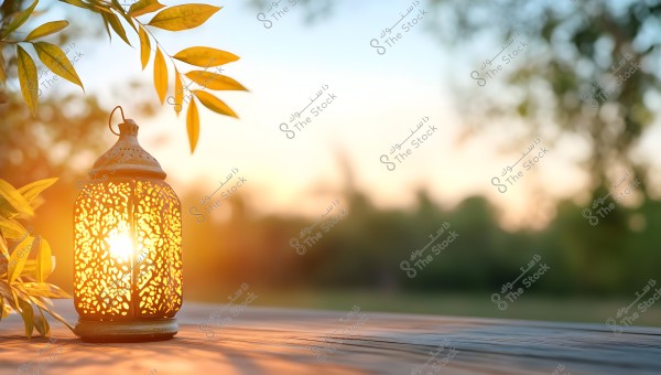 A intricately designed metal lantern illuminated, placed on a wooden outdoor table during sunset. Dangling green leaves from a tree add a natural touch, while the background shows a blurred forest with warm colors indicative of the day\'s end.