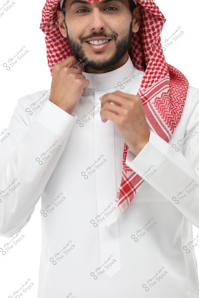 Image of a man wearing the traditional Saudi white thobe with a red and white checkered shemagh, offering a slight smile. The man is adjusting his attire, with a plain white background.