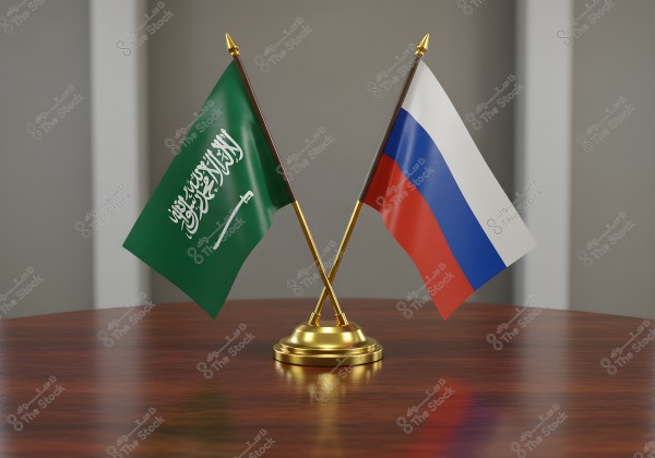 The image shows two crossed flags, with the flag of Saudi Arabia on the left featuring a green background with Arabic script and a white sword, and the flag of Russia on the right featuring three horizontal stripes in white, blue, and red. The flags are placed on a golden stand on a wooden table.