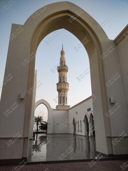 An image showcasing a view of a mosque with a prominent minaret in the center, framed by a large arch. The architectural design is strong with high white walls and a beautiful reflection on the glossy tiled floor. In the background, palm trees and greenery add a natural touch to the scene.